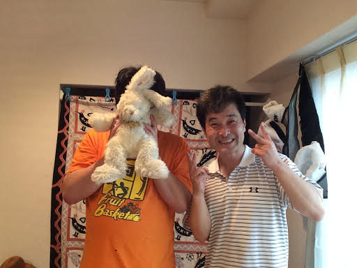 兎のぬいぐるみで顔を隠したオレンジのTシャツを着た大柄な男性と前田歩の笑顔のツーショット写真